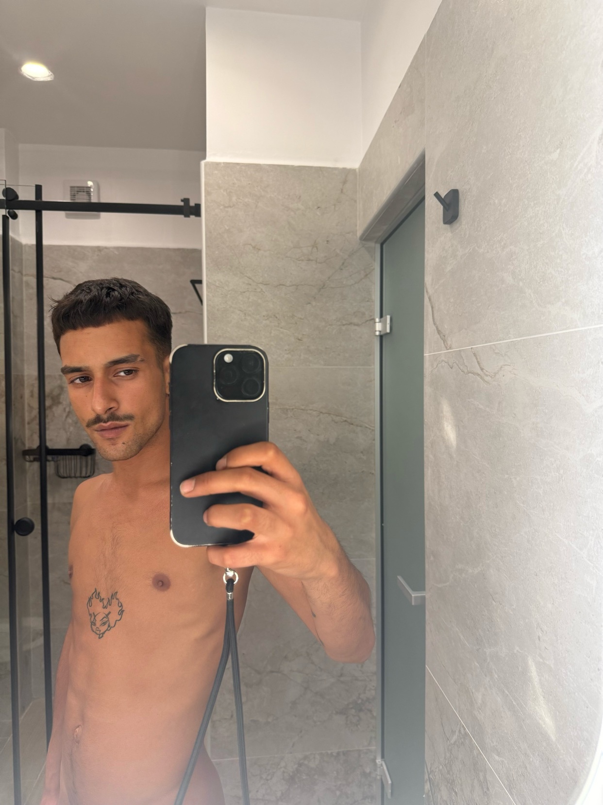 A man takes a mirror selfie in a bathroom. He is shirtless and has a mustache.