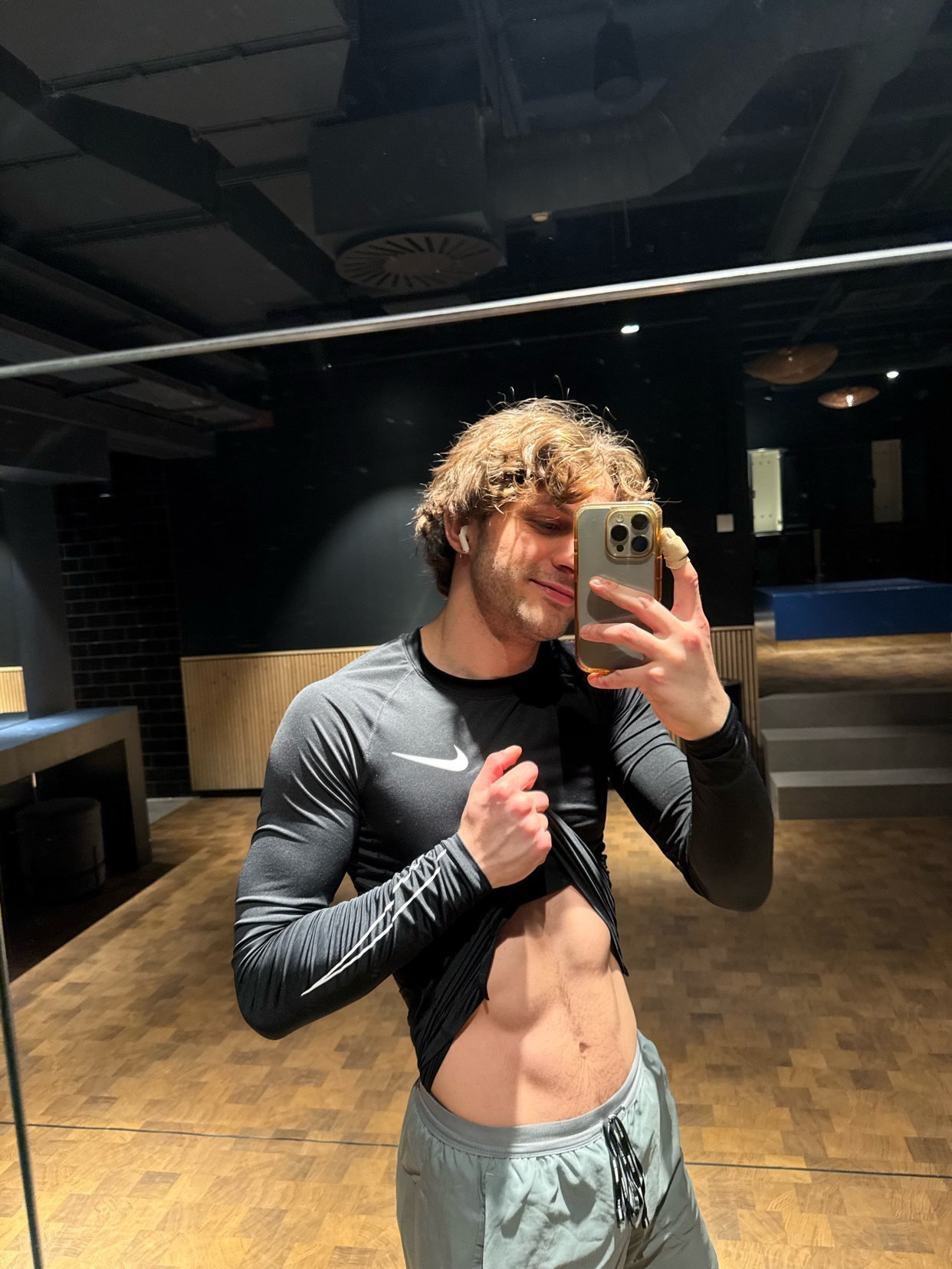 A man in a gym takes a mirror selfie, lifting his shirt to show his abs. He is wearing a long-sleeved black Nike shirt and grey sweatpants.