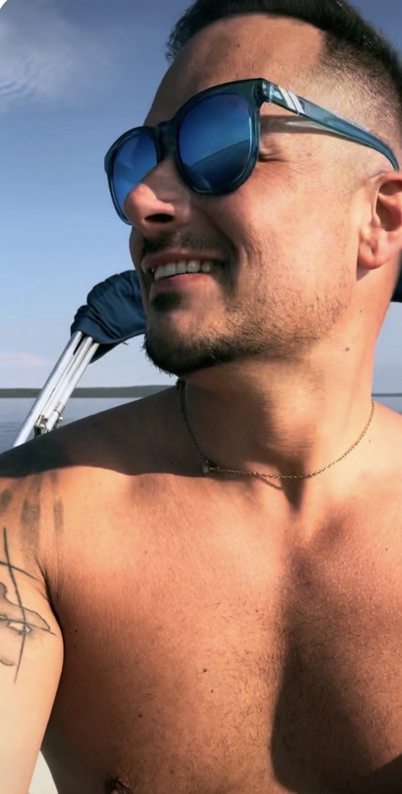A man with sunglasses and a tattoo on his arm smiles while looking out at the water on a sunny day.