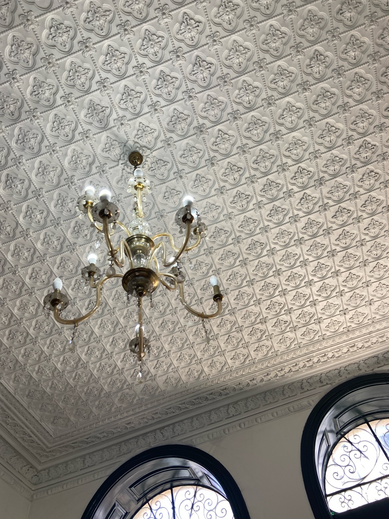 An ornate chandelier hangs from a highly decorative ceiling with a repeating floral pattern. Arched windows with intricate metalwork are visible below.