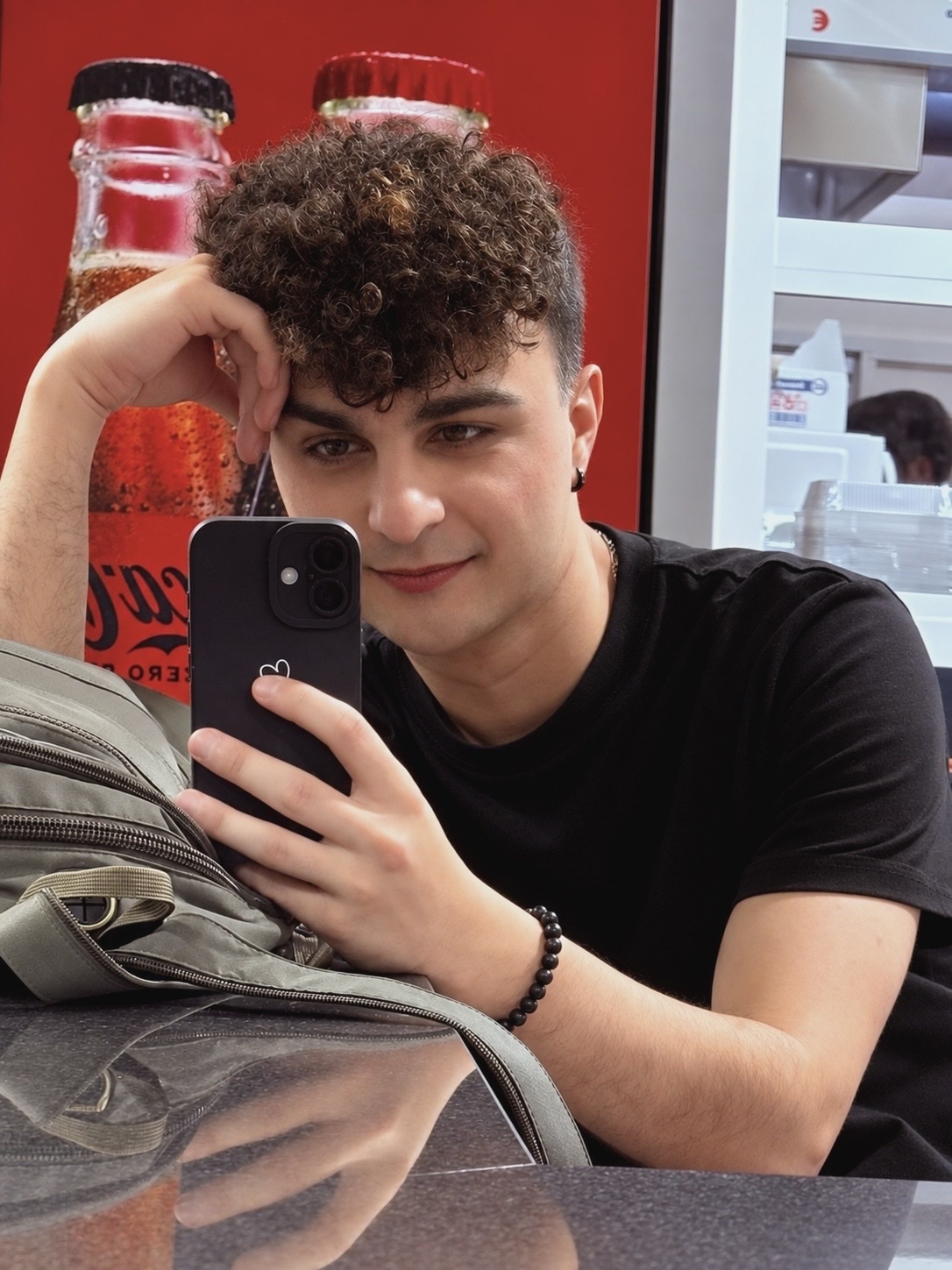 A young man with curly dark hair takes a selfie while leaning on a bag. He is wearing a black t-shirt and has a bracelet on his wrist. Behind him, two Coca-Cola bottles are visible.