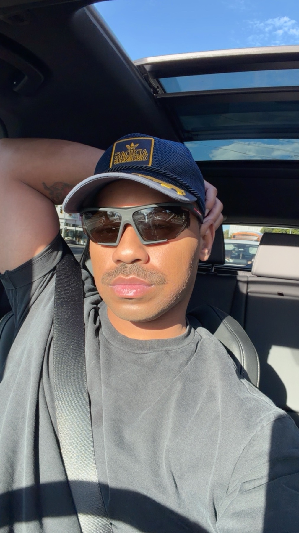 A man wearing sunglasses and a cap takes a selfie. He is shirtless.