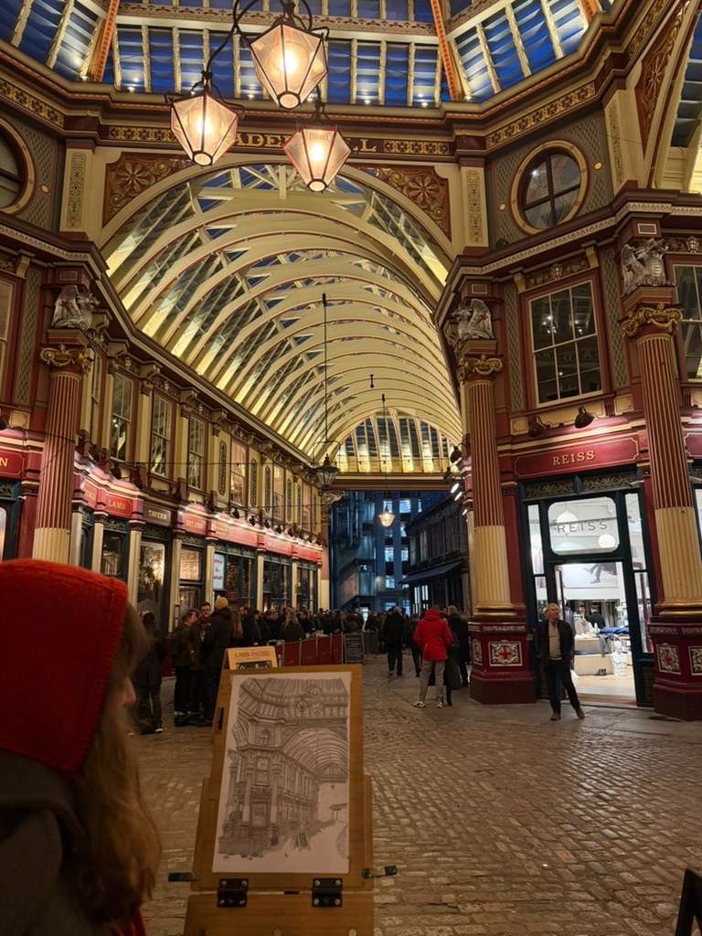 An ornate, historic market hall with a high, arched ceiling illuminated by decorative lamps. People stroll along the cobblestone floor, browsing shops, while a drawing of the interior is displayed on an easel in the foreground.