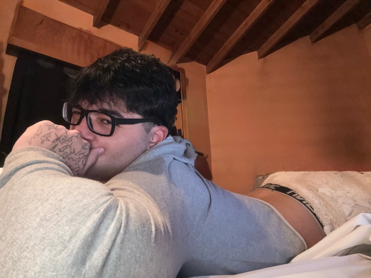 A man is lying on a bed, wearing glasses and a hoodie. He is shirtless and wearing underwear.