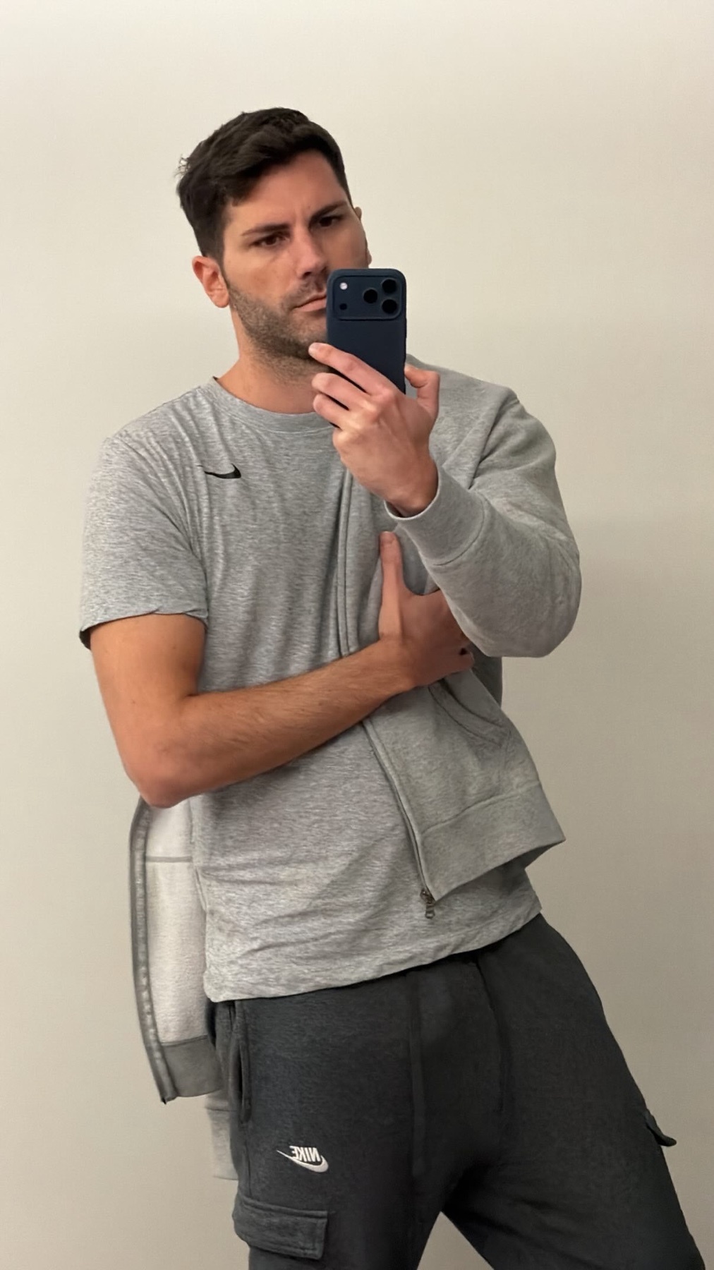 A man takes a selfie in a mirror, wearing a gray t-shirt and a gray sweatshirt. The man is looking at the camera.