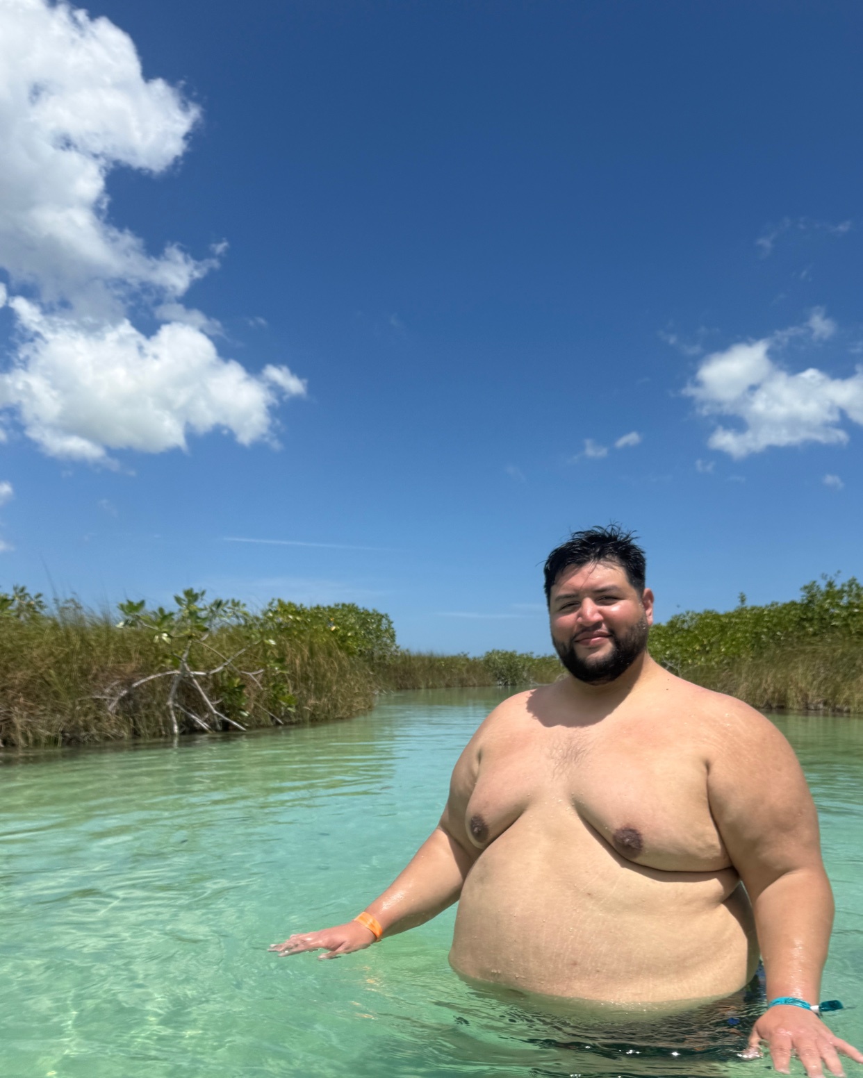 A shirtless man stands in clear turquoise water with lush green vegetation lining the banks under a bright blue sky with fluffy white clouds. The man is smiling and looking towards the camera.