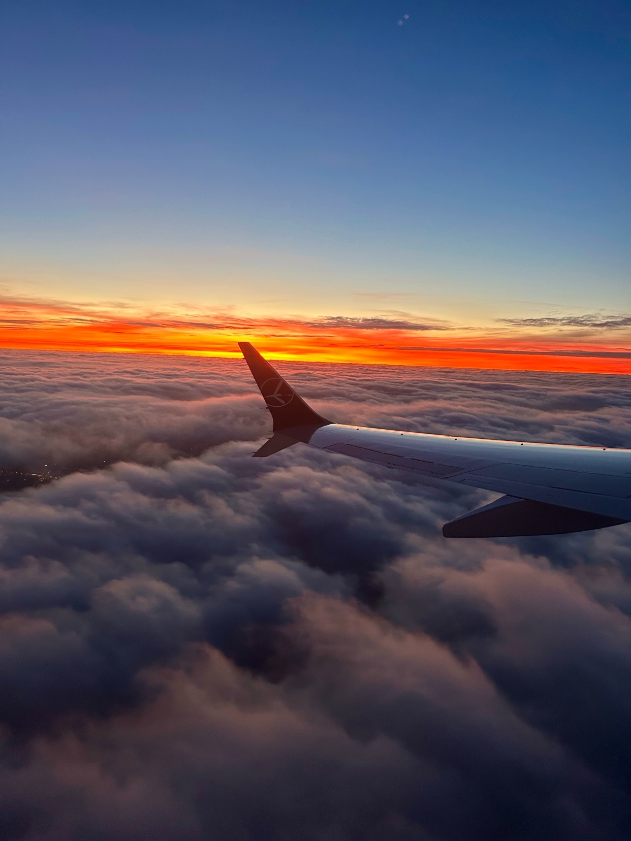 An airplane wing cuts through a sea of clouds at sunset. The sky is ablaze with orange and red hues.