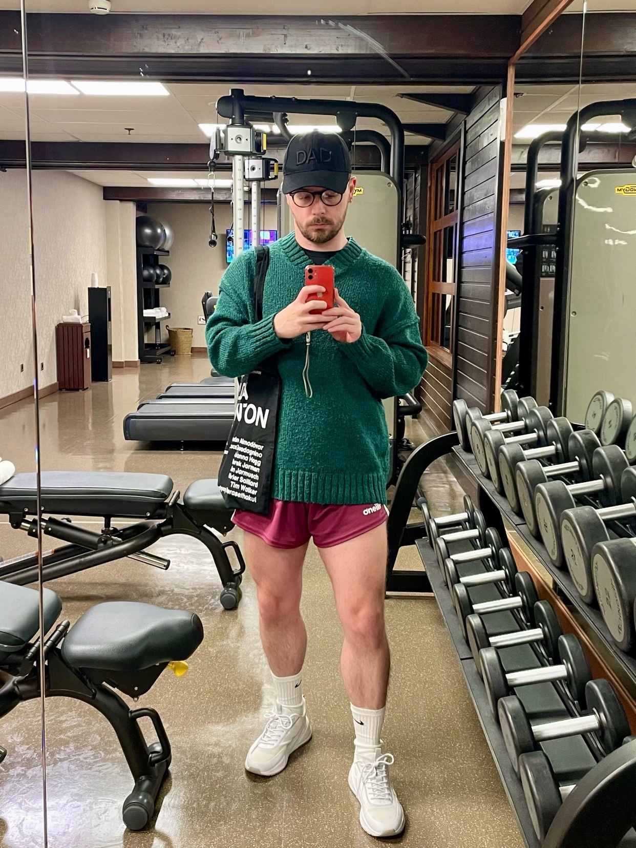 A man poses in a gym, taking a mirror selfie. He is wearing a green sweater, burgundy shorts, and white sneakers.