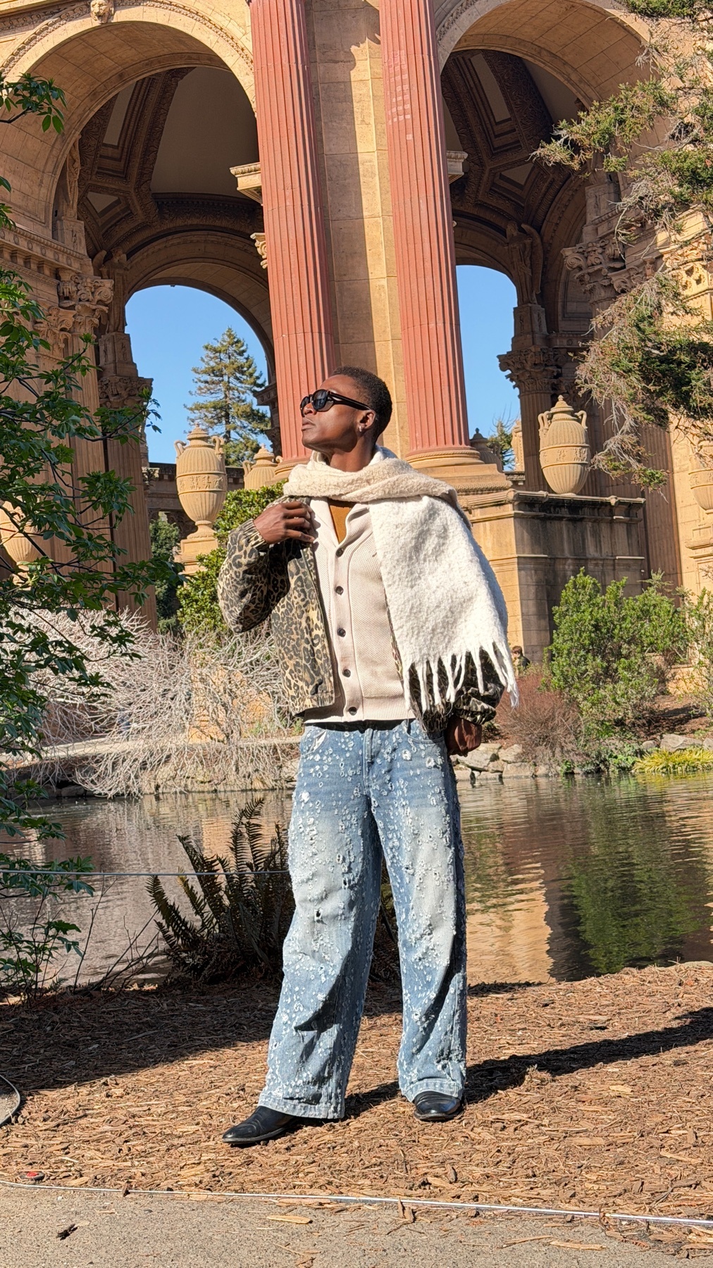 A man poses in front of the Palace of Fine Arts in San Francisco. He is wearing a stylish outfit with a jacket, scarf, and jeans.