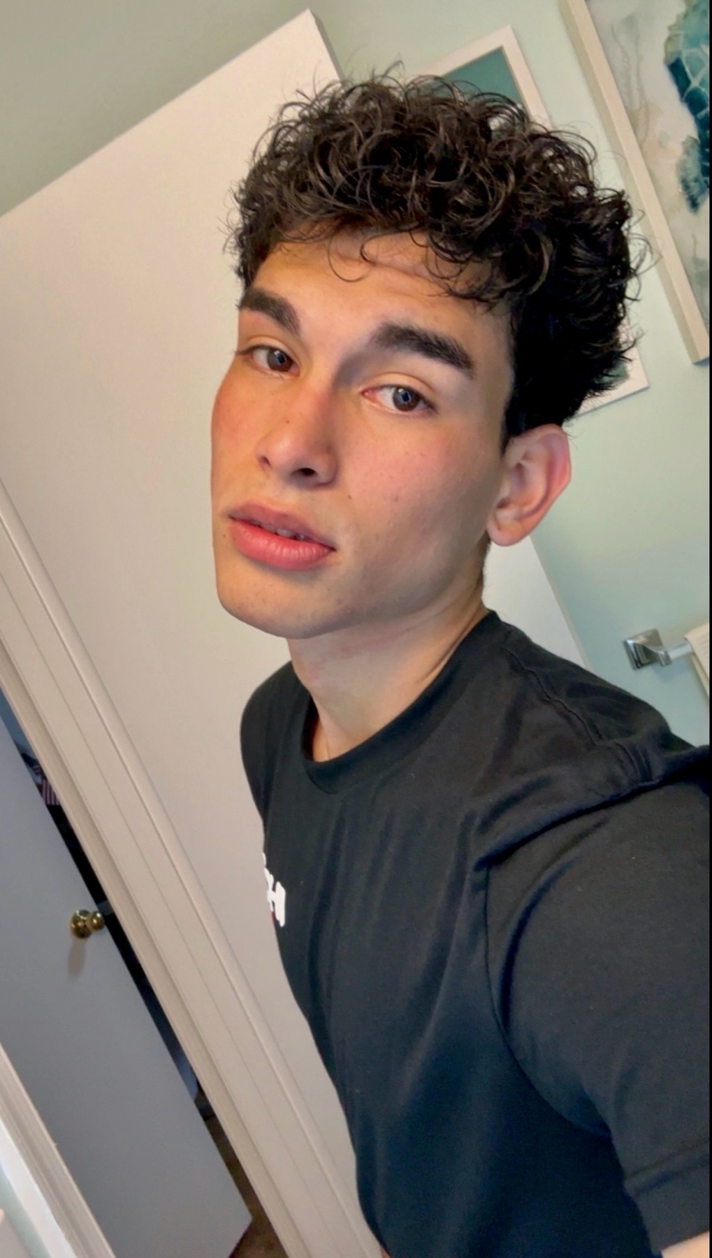 A close-up selfie of a young man with curly dark hair and a black t-shirt. He is looking towards the camera with a neutral expression.