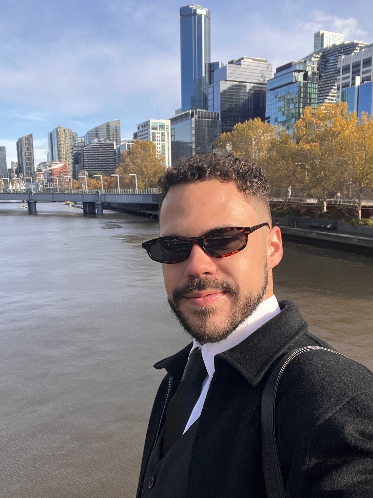 A man wearing sunglasses and a suit poses for a selfie with a cityscape and river in the background during autumn.