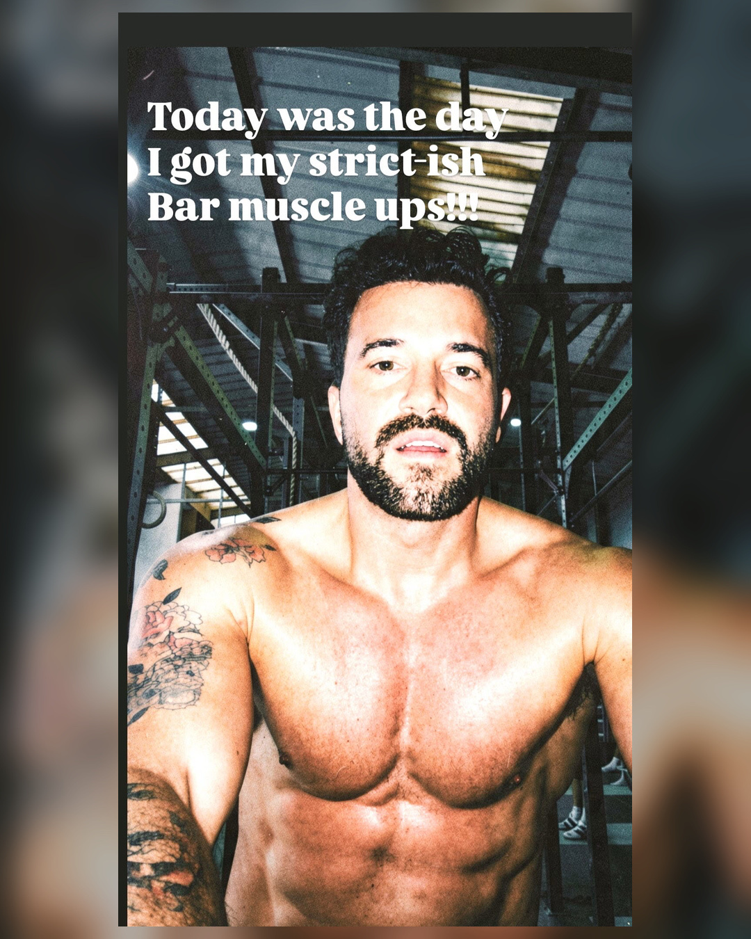 A shirtless man with tattoos on his arm poses for a selfie in a gym. He has a beard and short, dark hair, and his muscular chest and abs are prominent.