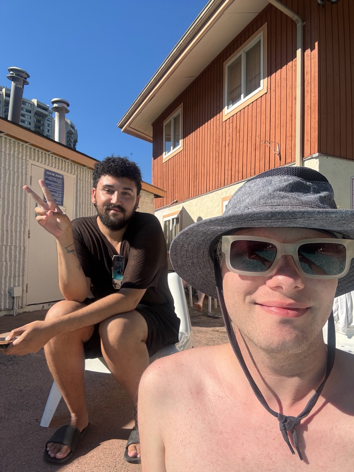 Two men smile for a selfie outdoors on a sunny day. The man in the foreground wears a hat and sunglasses, while the man in the background flashes a peace sign.