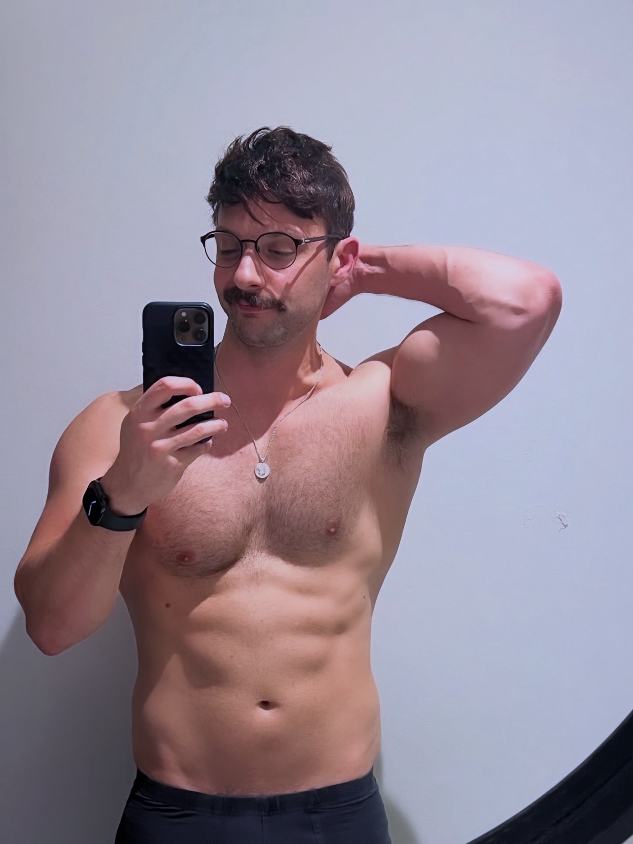 A man takes a mirror selfie, showcasing his physique. He is shirtless, with a mustache and glasses.