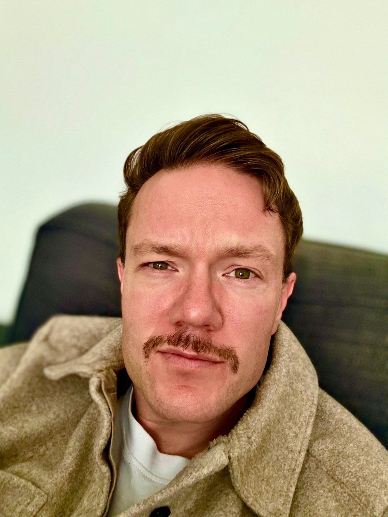 A man with a mustache and styled hair takes a selfie while wearing a tan jacket over a white t-shirt. He is looking directly at the camera.