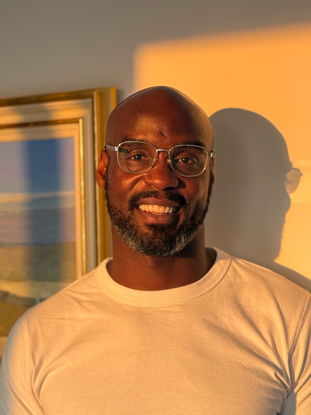 A portrait of a smiling Black man wearing glasses. He is bald with a beard and wearing a white shirt.