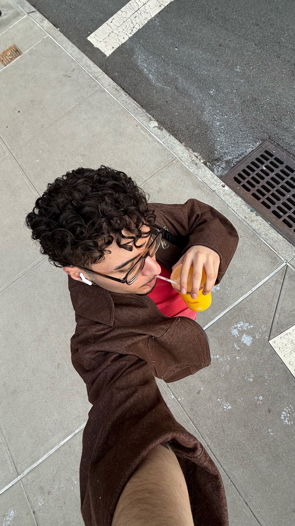 A man with curly hair and glasses takes a selfie while drinking from a cup on a sidewalk.