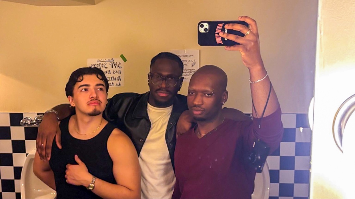 Three men pose for a selfie in what appears to be a public restroom. The man on the left wears a black tank top, the middle man wears a black leather jacket over a yellow shirt, and the man on the right wears a maroon v-neck shirt.