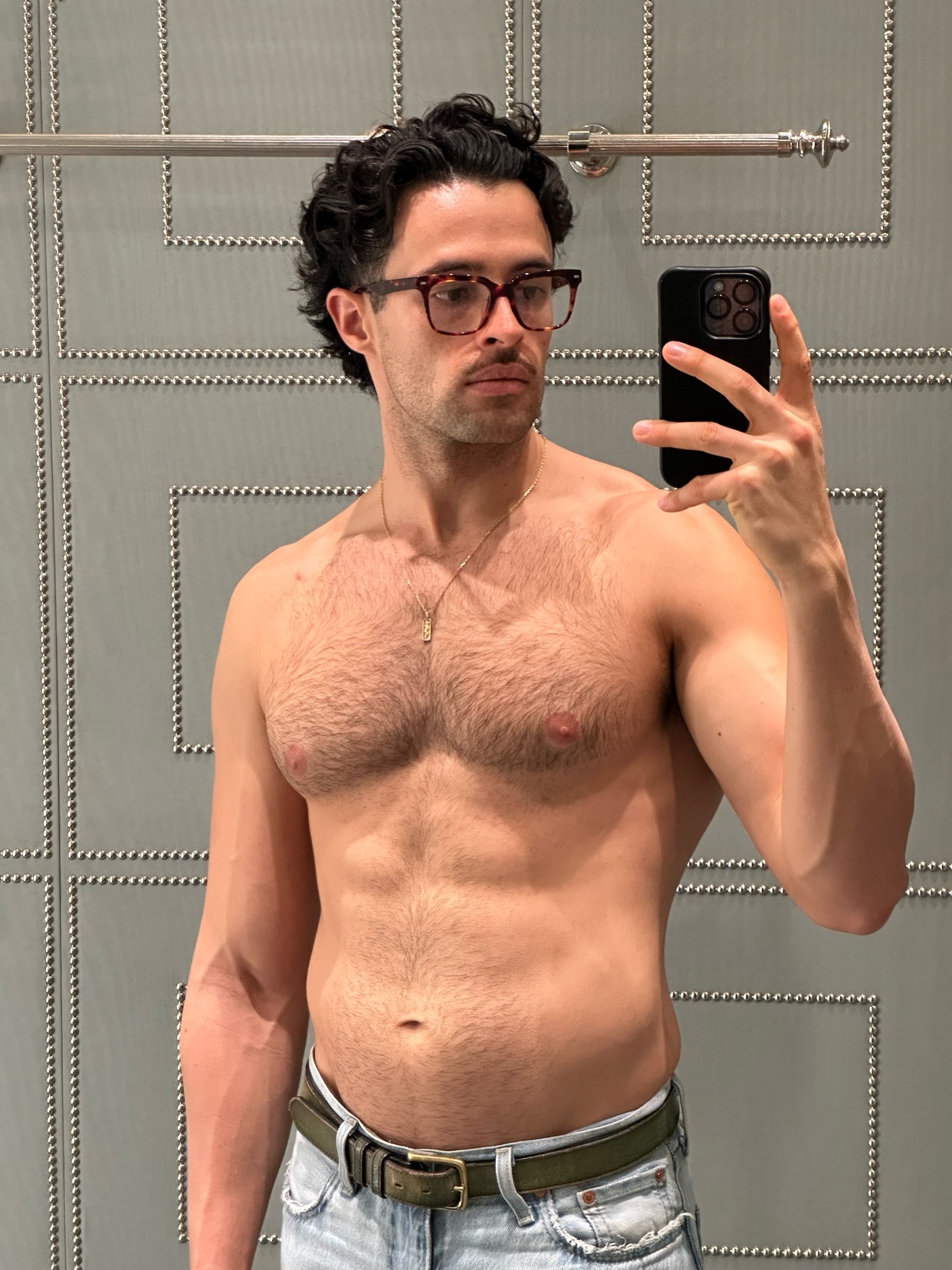 A man with dark curly hair and glasses takes a selfie in a changing room. He is shirtless, showing off his muscular physique.