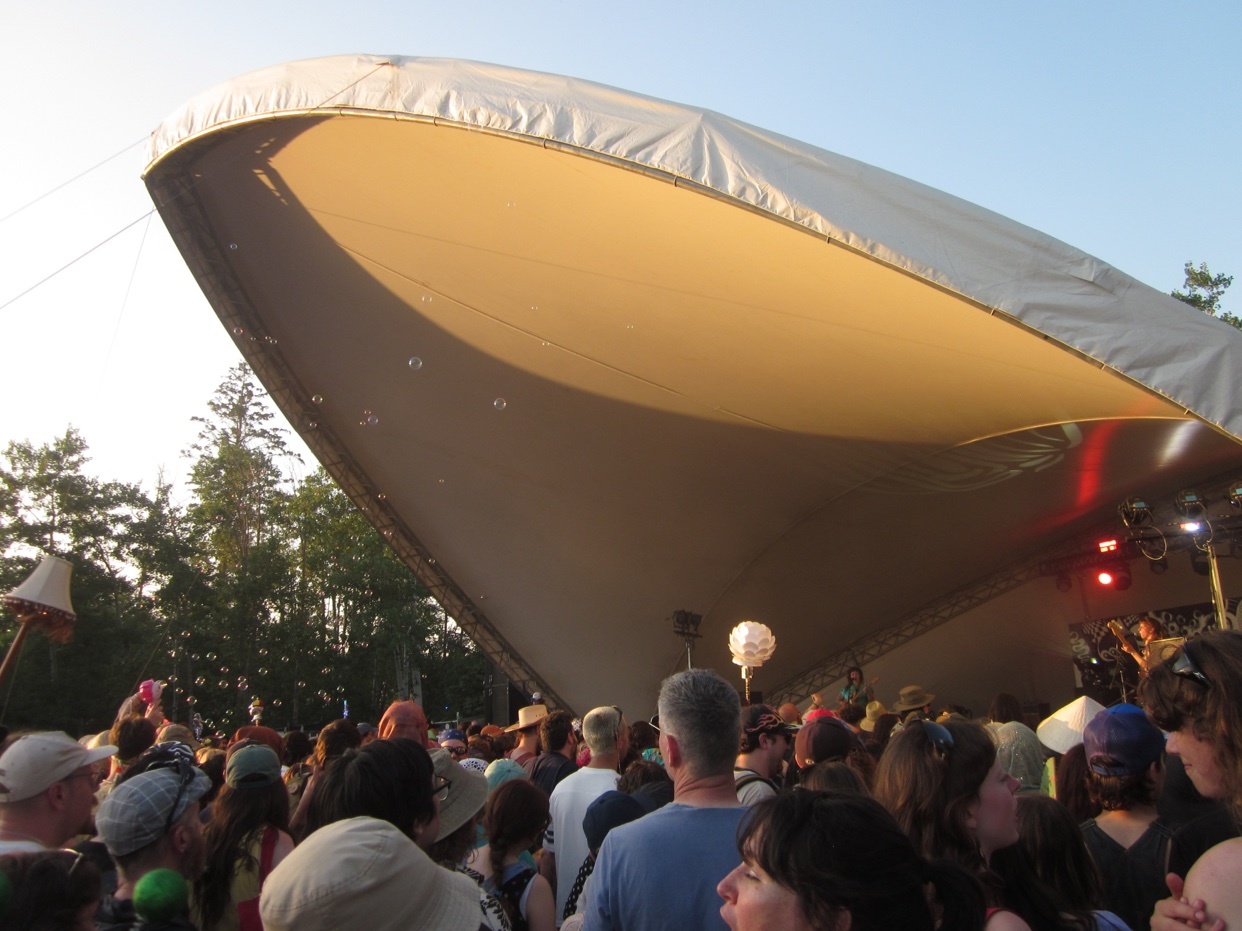 A large crowd gathers under a white tented stage at an outdoor concert. The sun is setting, casting a warm glow on the scene.