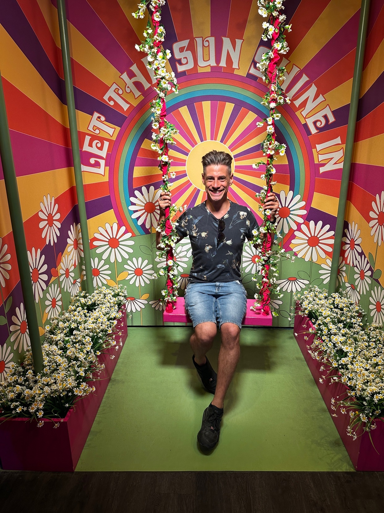 A man smiles while sitting on a swing in front of a colorful backdrop. The backdrop features a sun design and the words "Let the sun shine in."