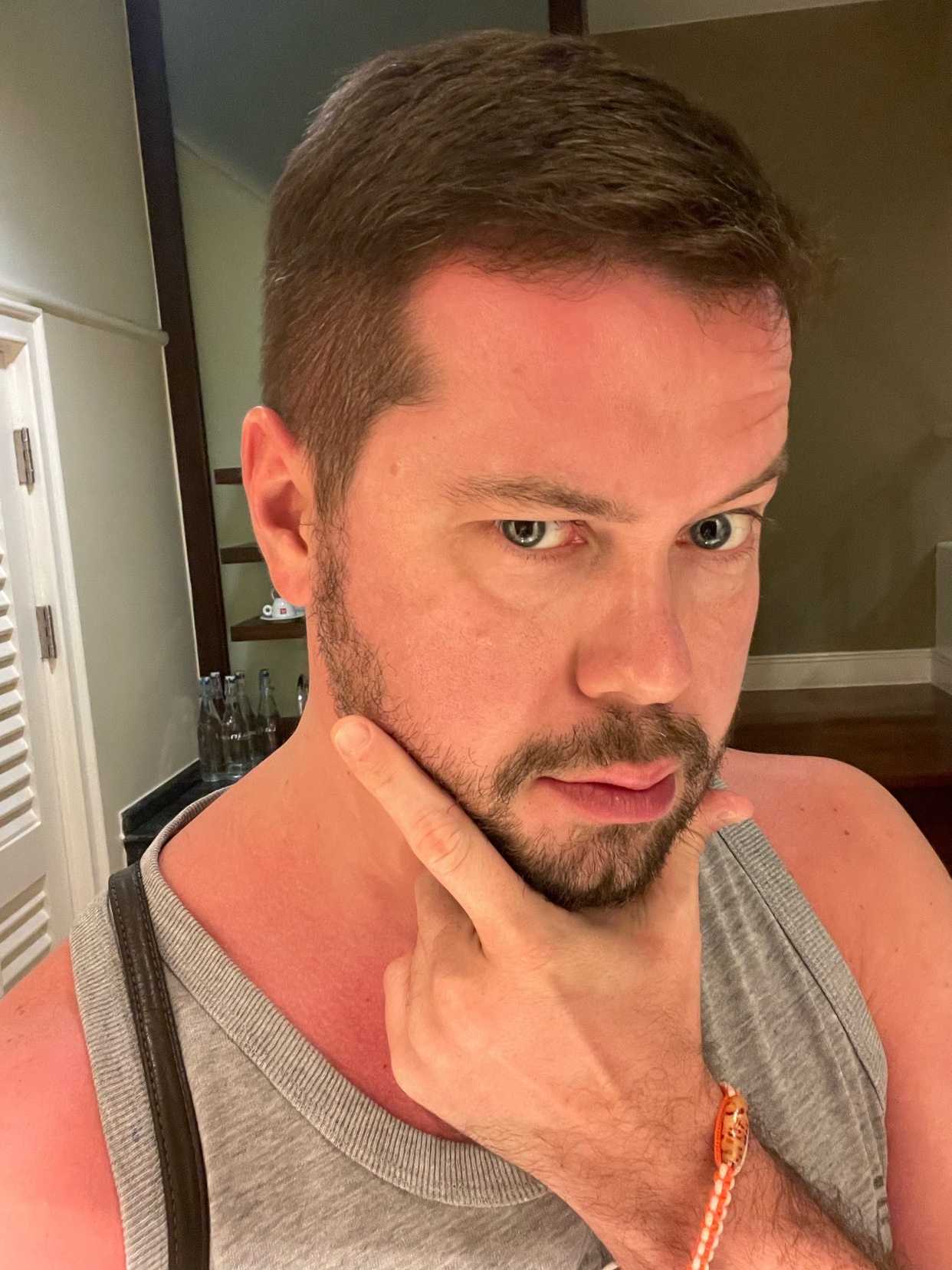 A man takes a selfie, looking directly at the camera. He is shirtless and has a tan.