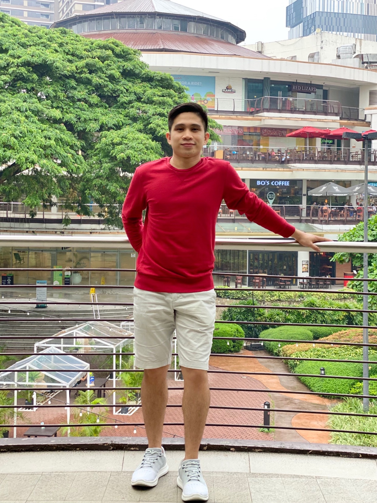 A man poses for a photo outdoors. He is wearing a red long-sleeved shirt and white shorts.