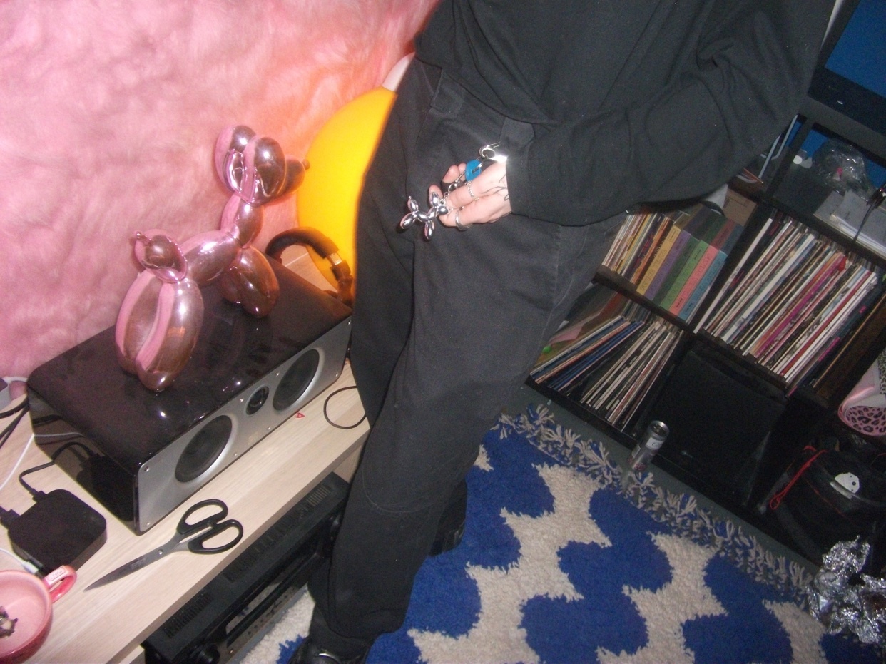 A shiny pink balloon dog sits on top of a speaker next to a stack of records. The image is taken from a low angle, showing a person in black pants and a black top.