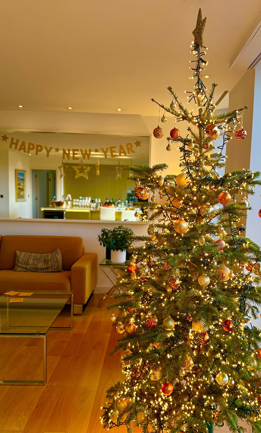 A decorated Christmas tree is the focal point of a living room, with a "Happy New Year" banner in the background. The room is warmly lit, creating a festive atmosphere.