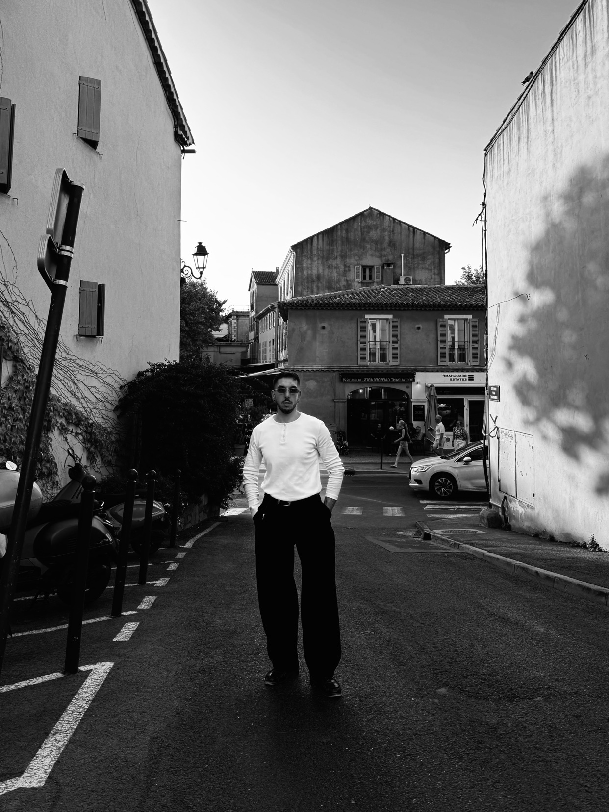 A man stands in the middle of a narrow street in St. Tropez, France. He is wearing a long-sleeved shirt, dark pants, and sunglasses. The black and white photo captures the European architecture of the surrounding buildings.
