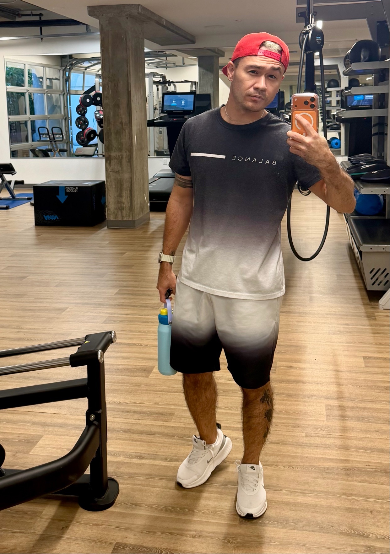 A man takes a mirror selfie at the gym. He is wearing a t-shirt, shorts, and sneakers.