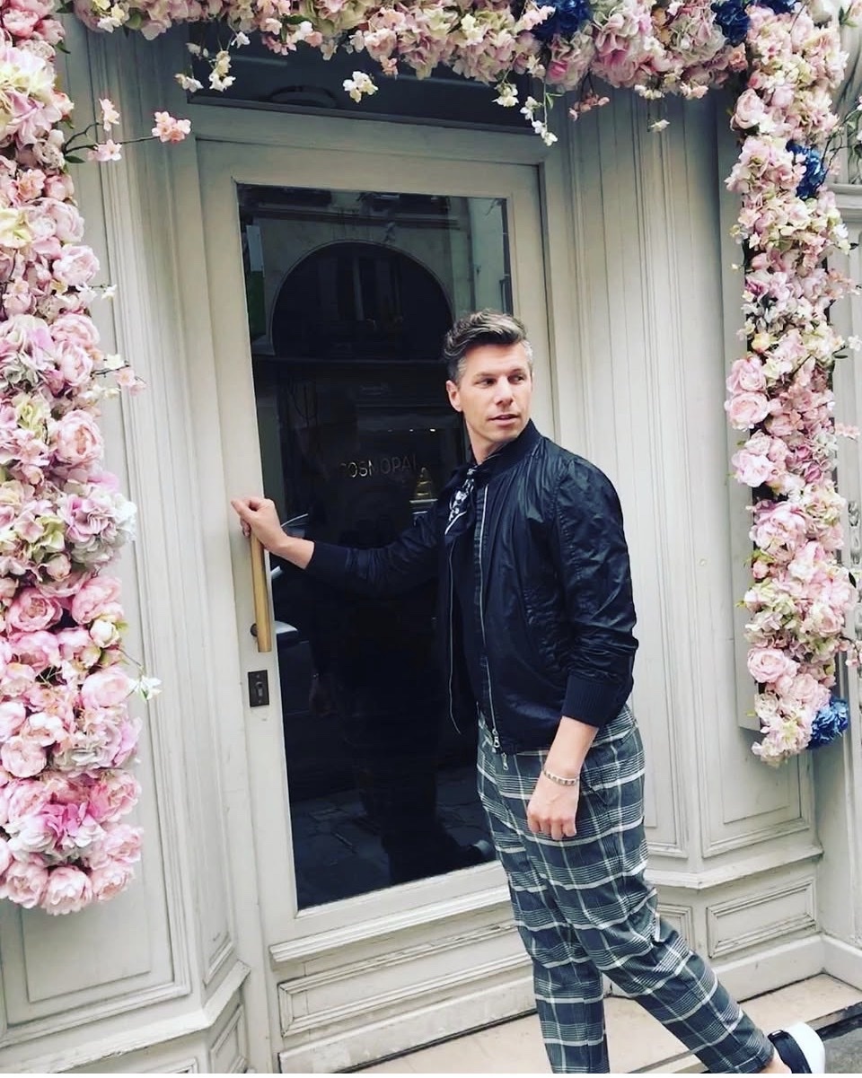 A man stands in front of a doorway adorned with floral arrangements. He is wearing a black bomber jacket, a black t-shirt, and plaid pants. The man is looking to his left and has one hand on the door handle.