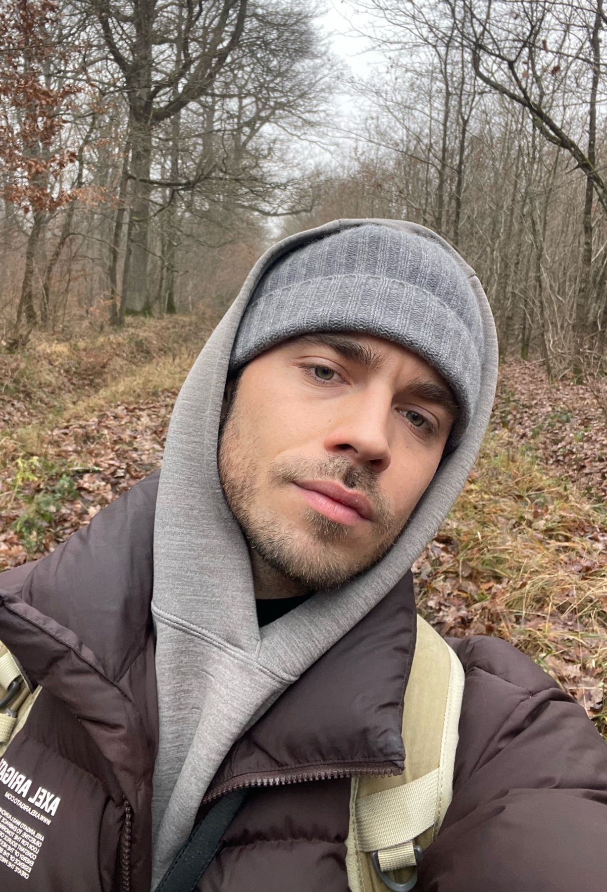 A man takes a selfie while hiking in a forest. He is wearing a beanie and a hoodie.