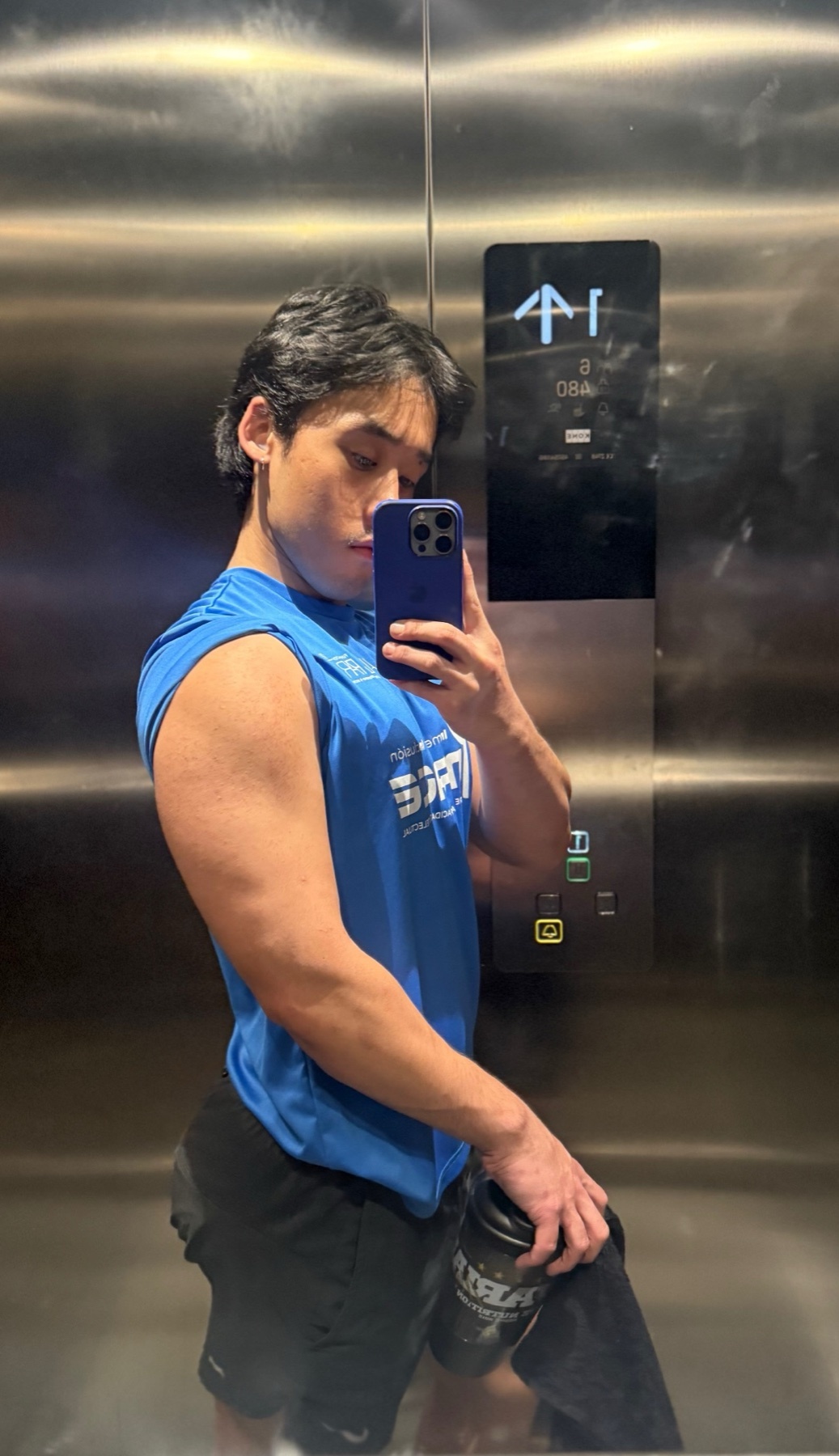 A man in a blue sleeveless shirt takes a selfie in an elevator, flexing his bicep. He is holding a shaker bottle and a towel.