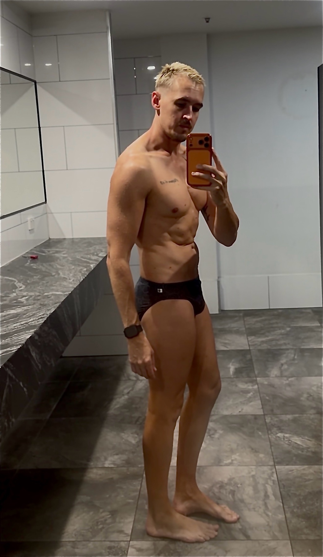 A man with blonde hair and tattoos poses for a selfie in a gym locker room, wearing swim briefs and showing off his muscular physique.