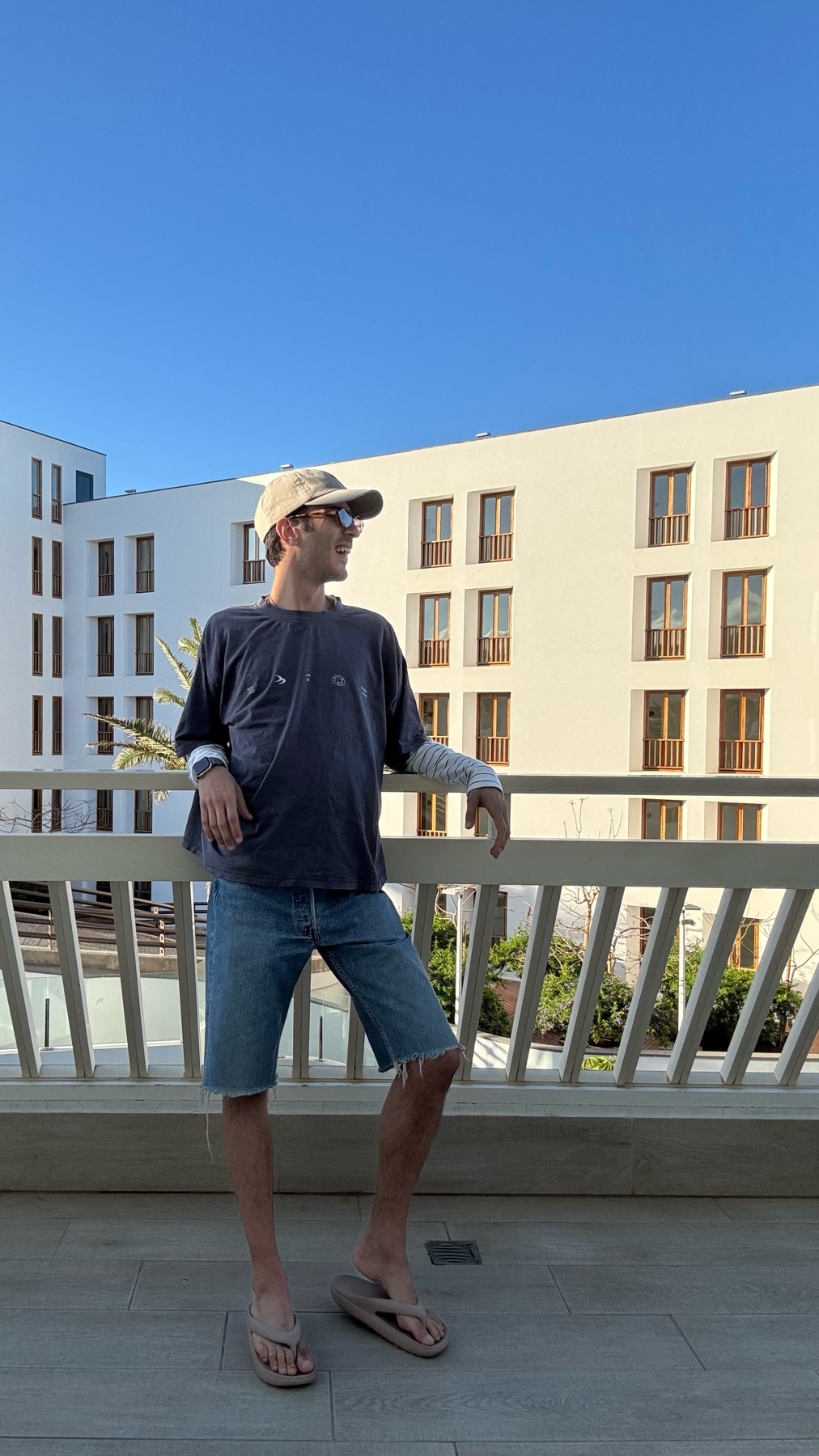 A man stands on a balcony wearing a t-shirt, denim shorts, a baseball cap, and flip-flops. He is leaning on a railing with a modern building in the background.