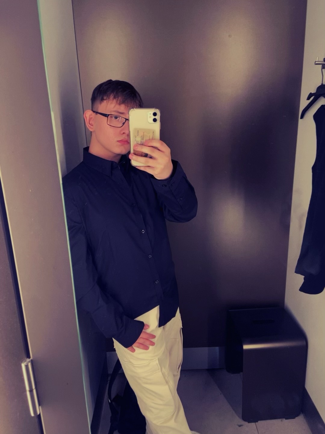 A man takes a mirror selfie in a fitting room. He is wearing glasses, a dark shirt, and beige pants.