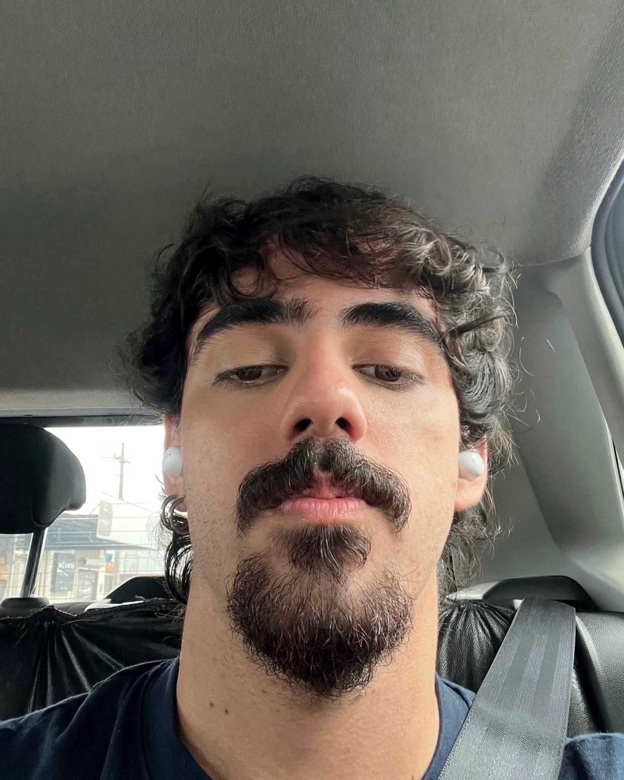 A selfie of a man with curly hair, a beard, and a mustache. He is wearing earbuds and looking at the camera.