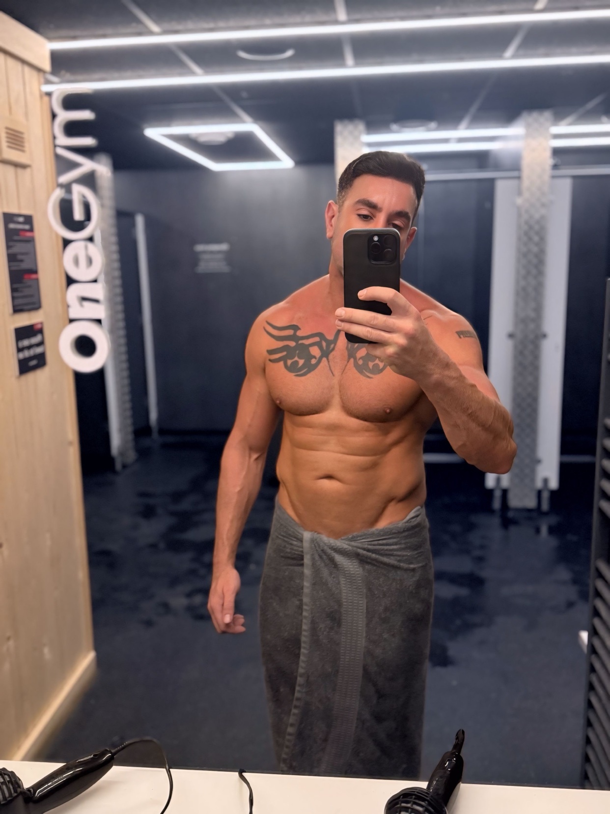 A man takes a mirror selfie in a gym locker room. He is shirtless, with a towel wrapped around his waist, and has visible tattoos.