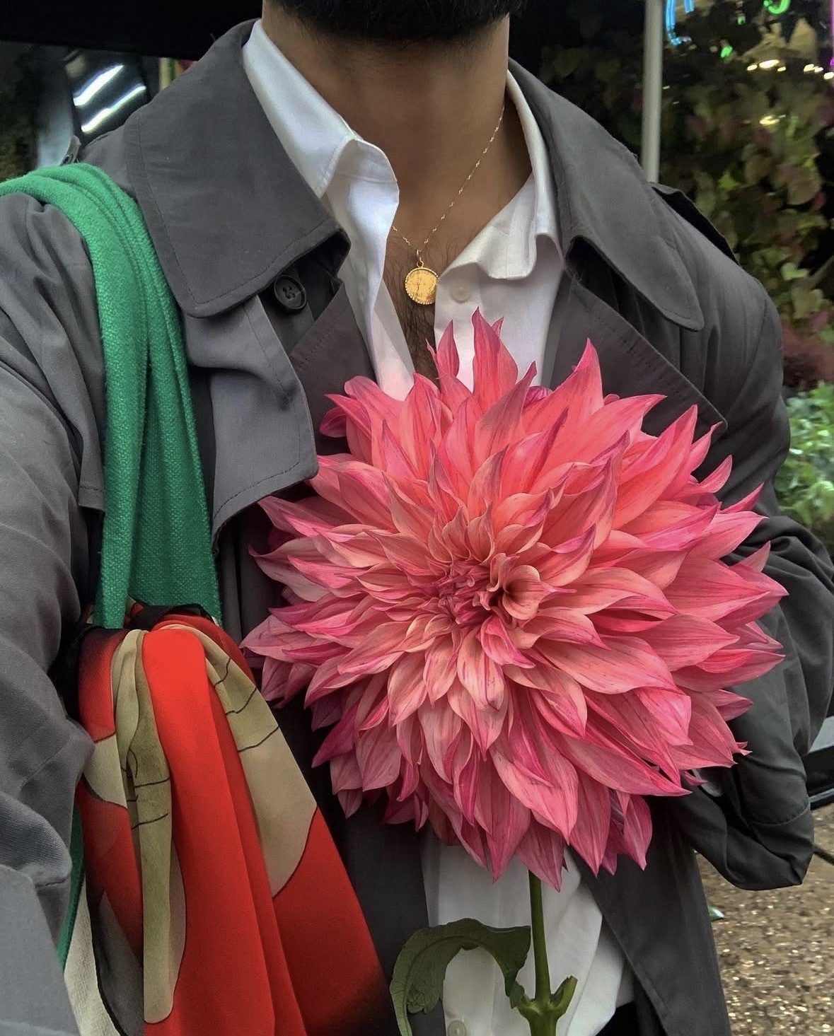 A man is pictured from the chest up, holding a large, vibrant pink dahlia flower. He is wearing a white button-down shirt, a grey trench coat, and a gold necklace.