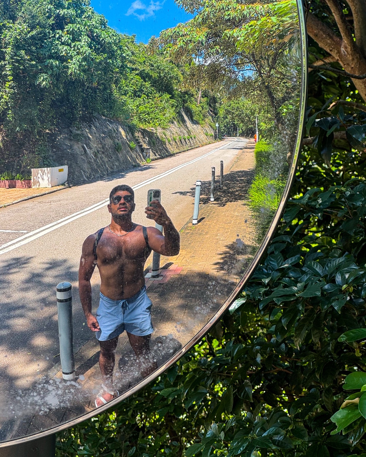 A man takes a shirtless selfie in a mirror outdoors. The photo captures his muscular physique and the scenic road behind him.