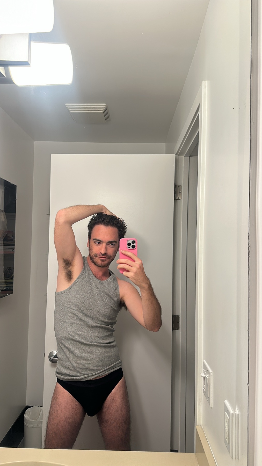 A man in a grey tank top and black underwear poses for a mirror selfie in a bathroom.
