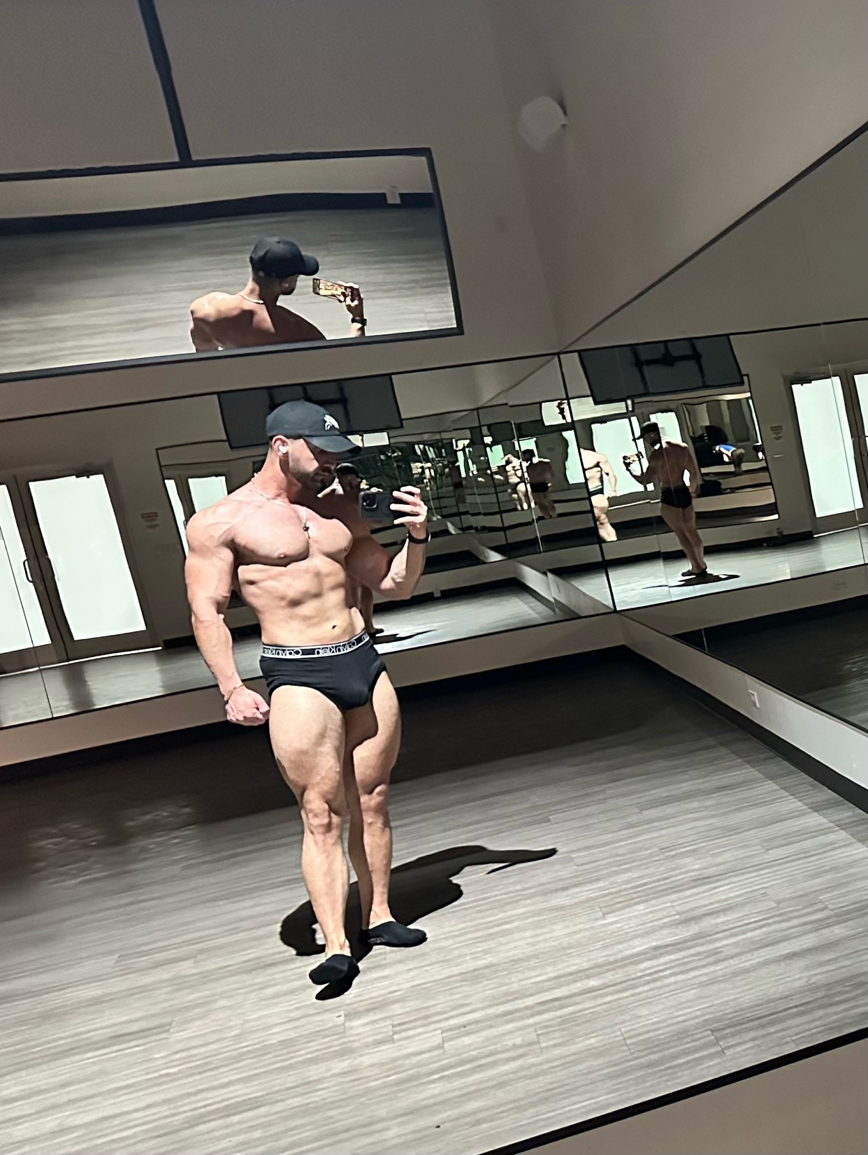 A muscular man takes a mirror selfie at the gym. He is shirtless and wearing black underwear.