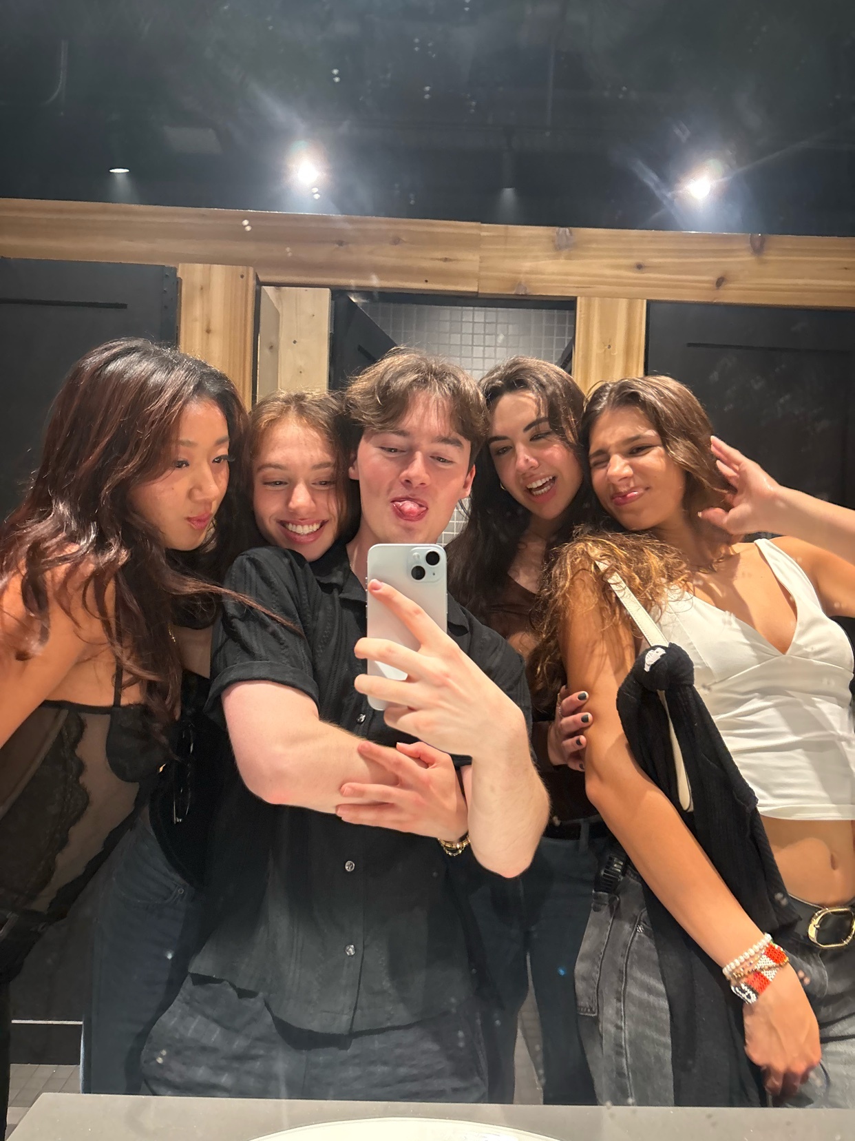 A group of friends take a selfie in a bathroom mirror. They are smiling and posing for the camera, enjoying a night out.