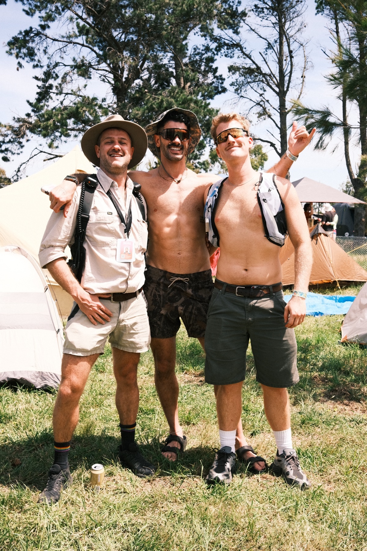 Three men pose for a photo outdoors, two of them shirtless. They are smiling and appear to be enjoying themselves.