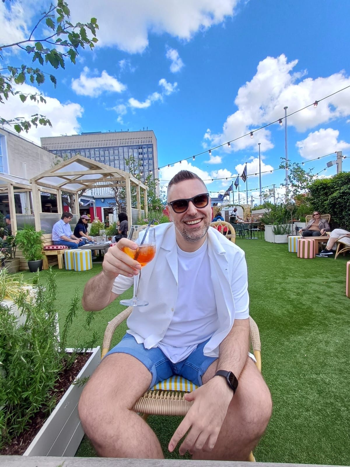 A man in sunglasses and a white shirt smiles while holding a cocktail in an outdoor seating area.