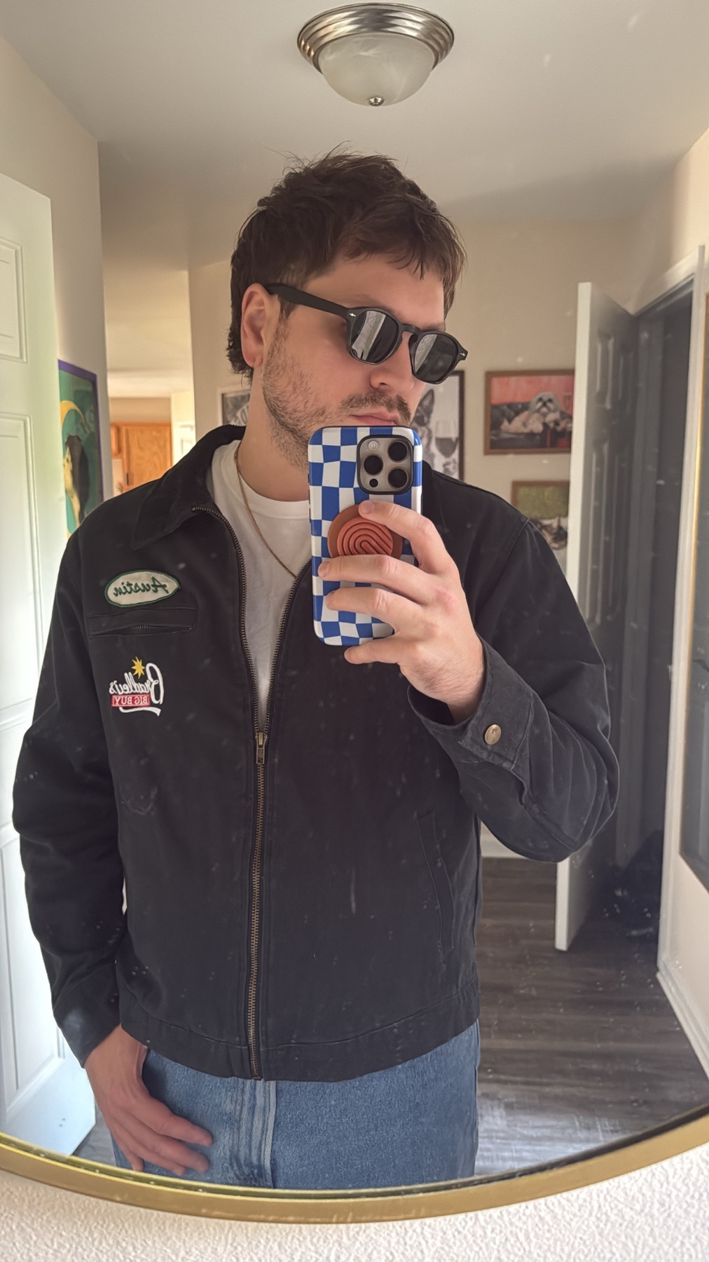A man with a mullet and sunglasses takes a mirror selfie in a black jacket and jeans.