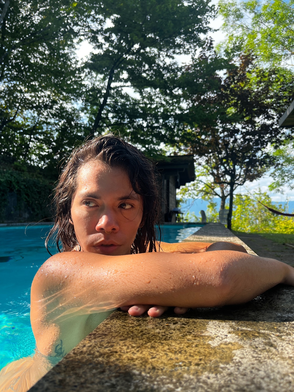 A man with long, dark hair rests his arm on the edge of a pool, looking off to the side. The sun shines brightly, illuminating the trees and water around him.