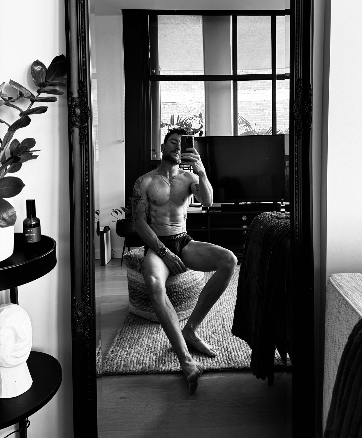 A man with tattoos and a muscular physique takes a mirror selfie while wearing underwear. He is sitting in a room with plants and a television in the background.
