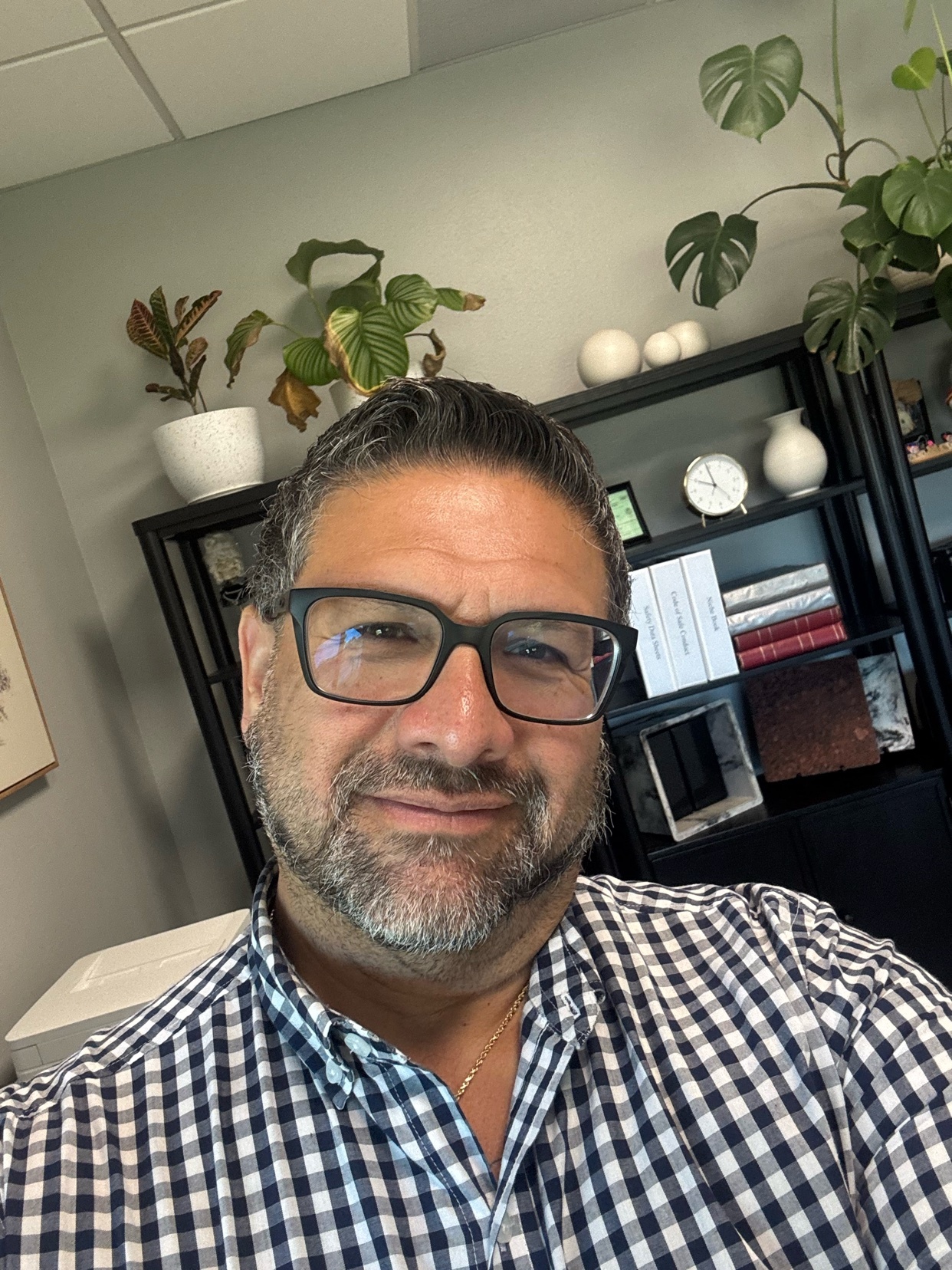 A man with glasses and a beard smiles at the camera, wearing a blue and white plaid shirt. He is in an office setting with plants and a bookshelf in the background.
